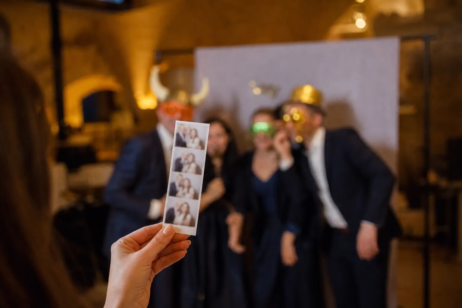 Roka drži natisnjen trak fotografij, v ozadju gostje z rekviziti pozirajo pred ozadjem foto kabine